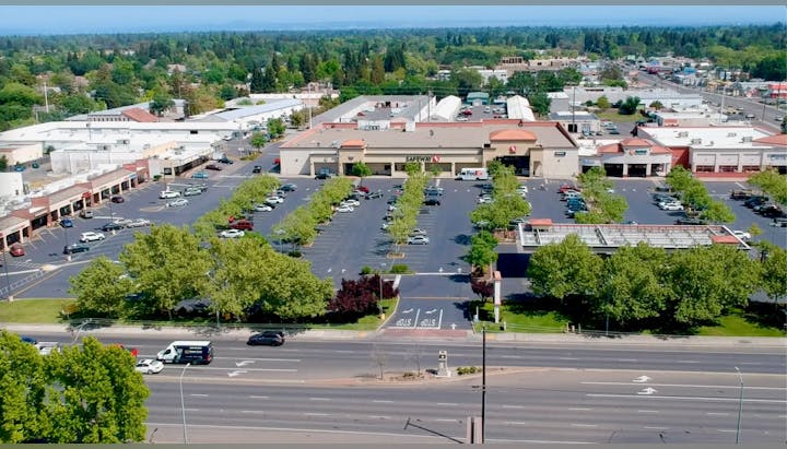 Aerial view of a shopping center with parking lot, various storefronts including a Safeway, and surrounding roads with vehicles.