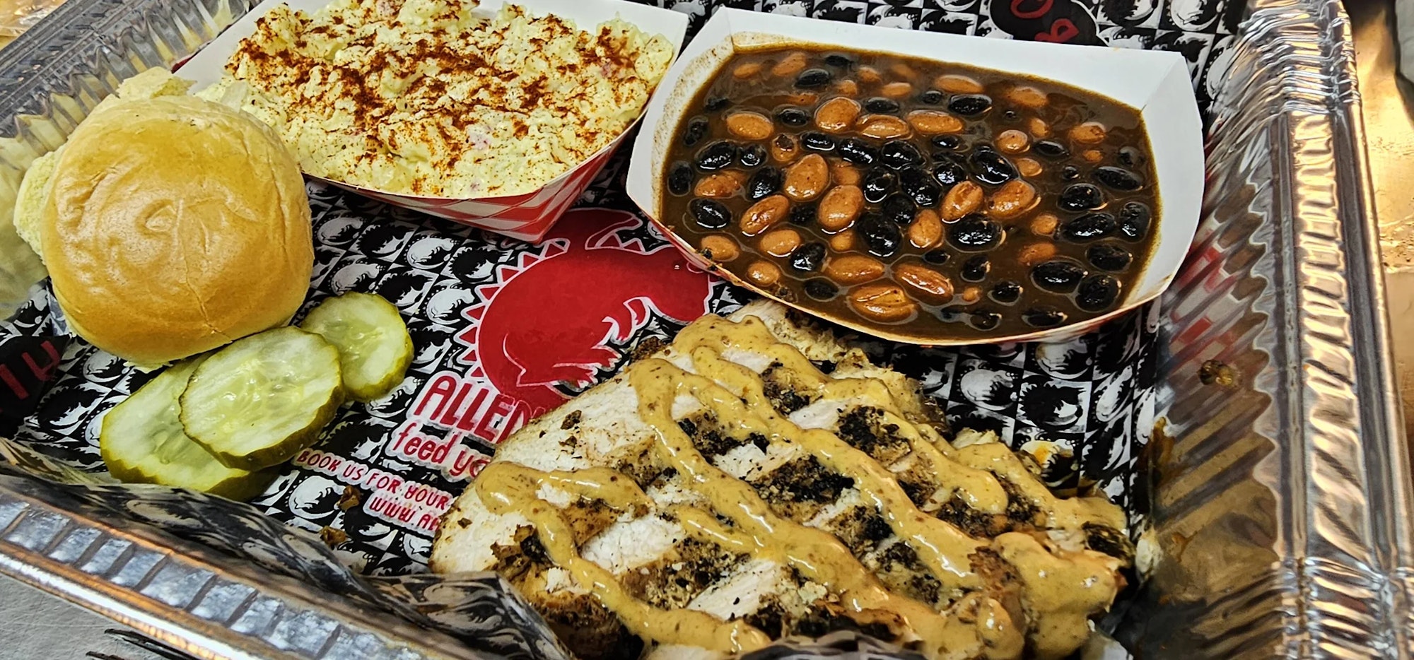 Grilled chicken with sauce, sliced pickles, a roll, potato salad, and beans in a tray.