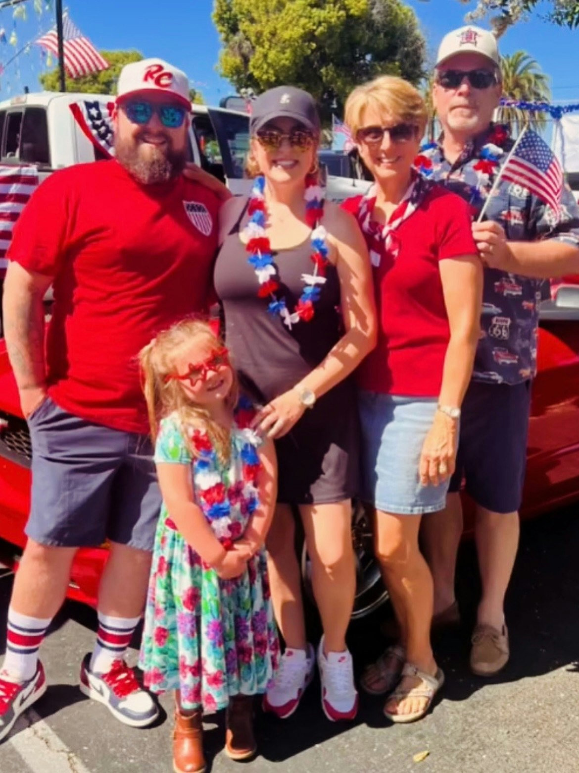 A group of people dressed in red, white, and blue, with American flags, standing in front of a vehicle.