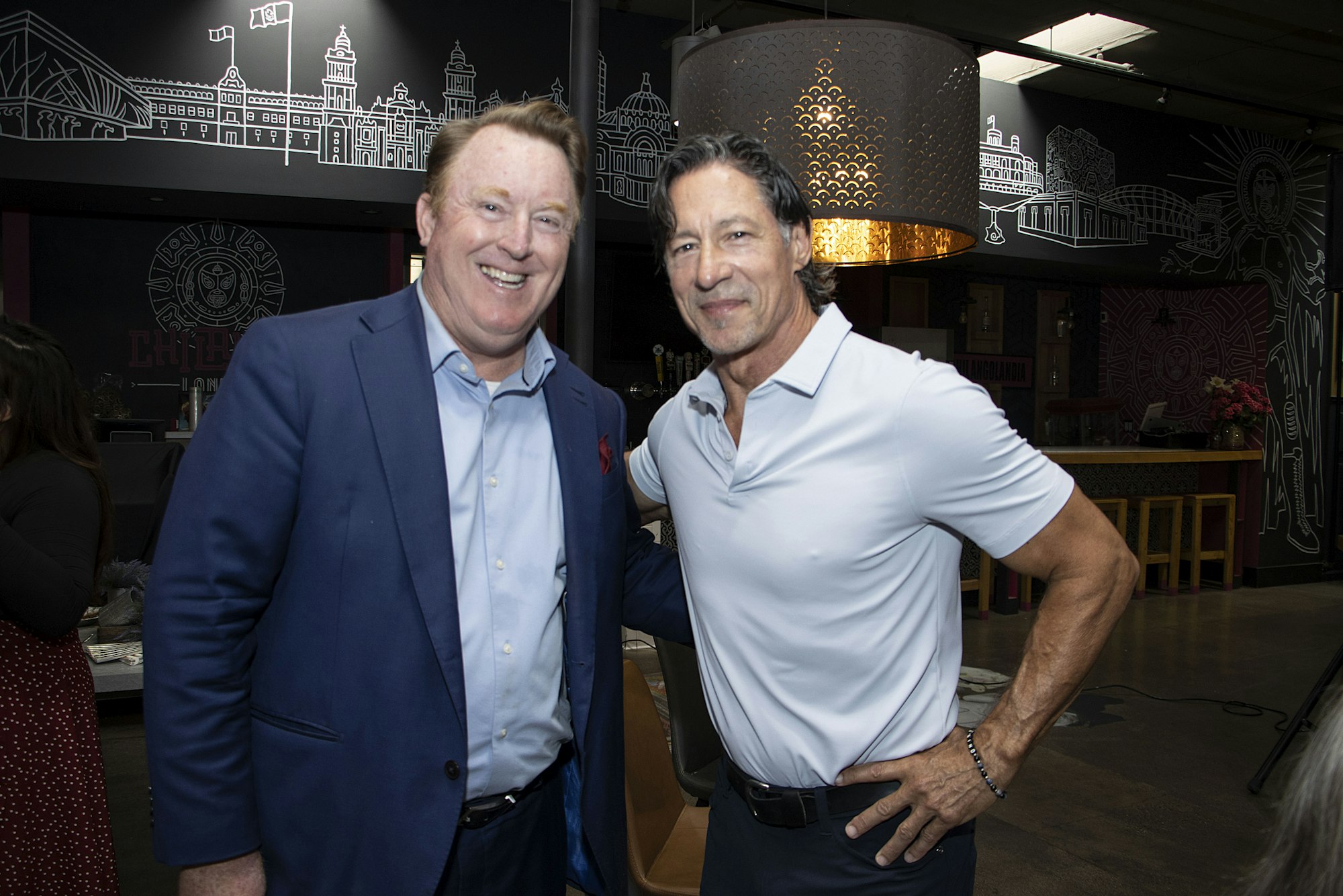 Two men smiling and posing together indoors, one in a suit and the other in a light shirt.