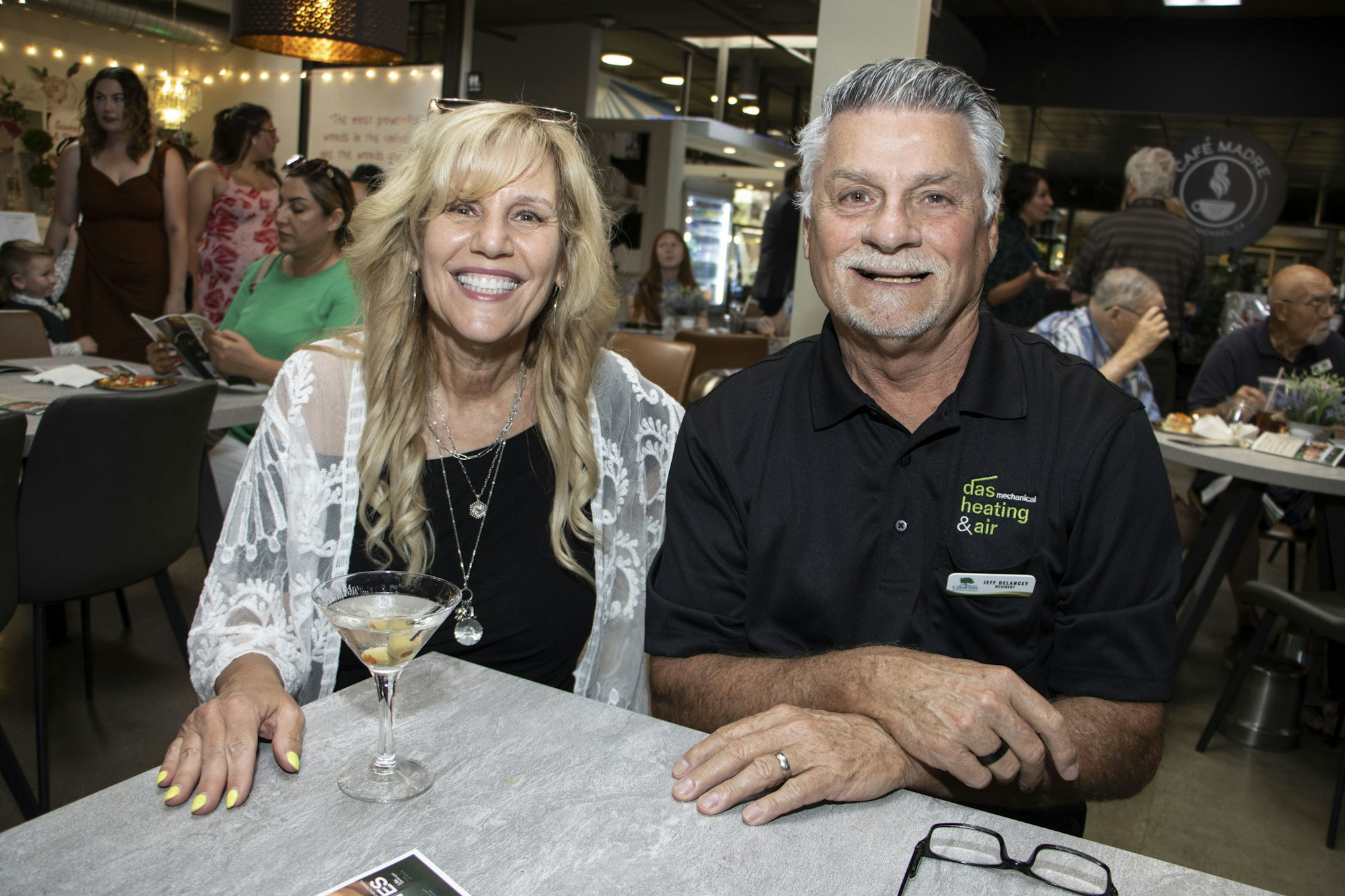 A smiling couple sits at a table in a lively restaurant with drinks and other people around.