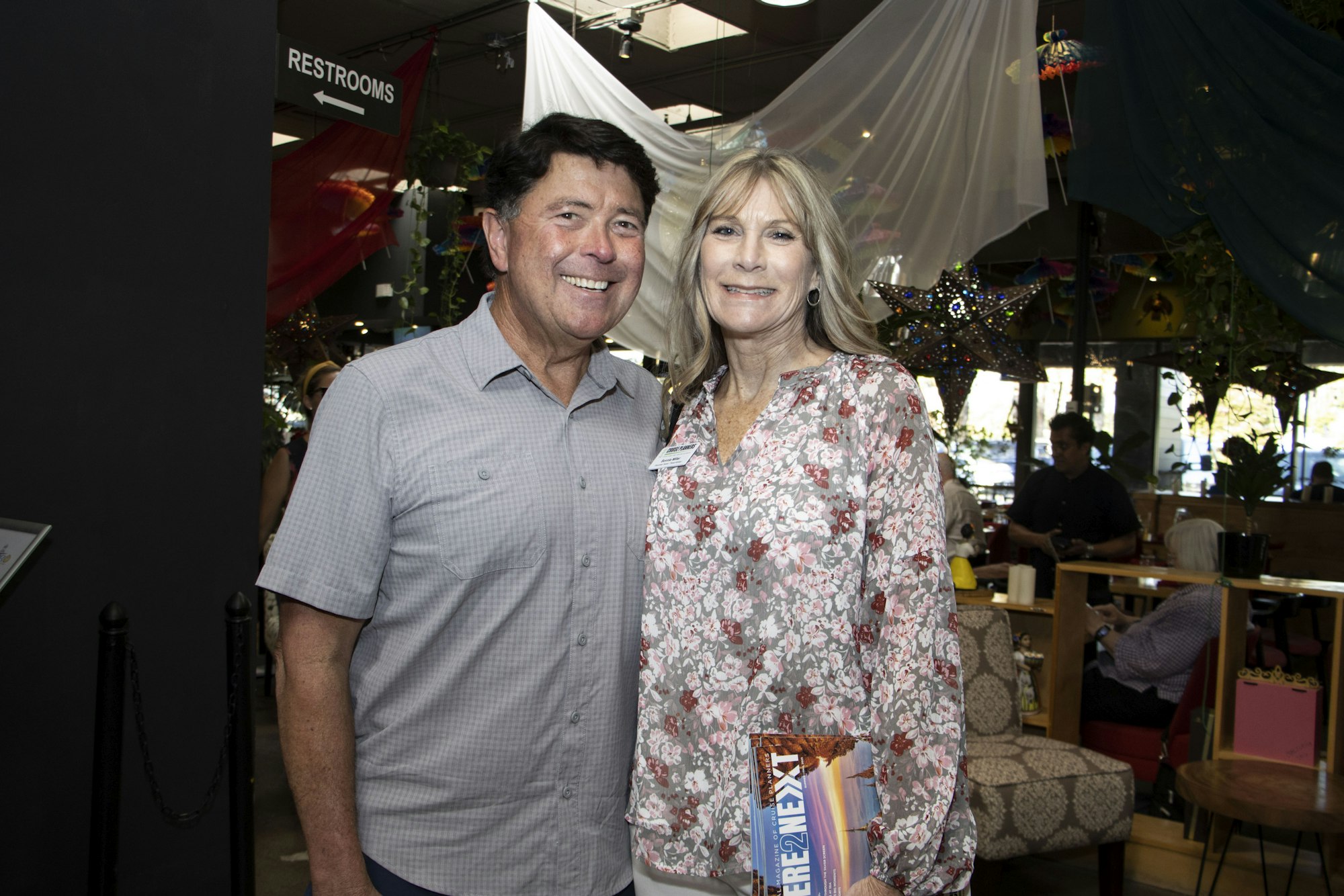A man and woman smiling, standing together indoors under colorful decor, with "RESTROOMS" sign above them.