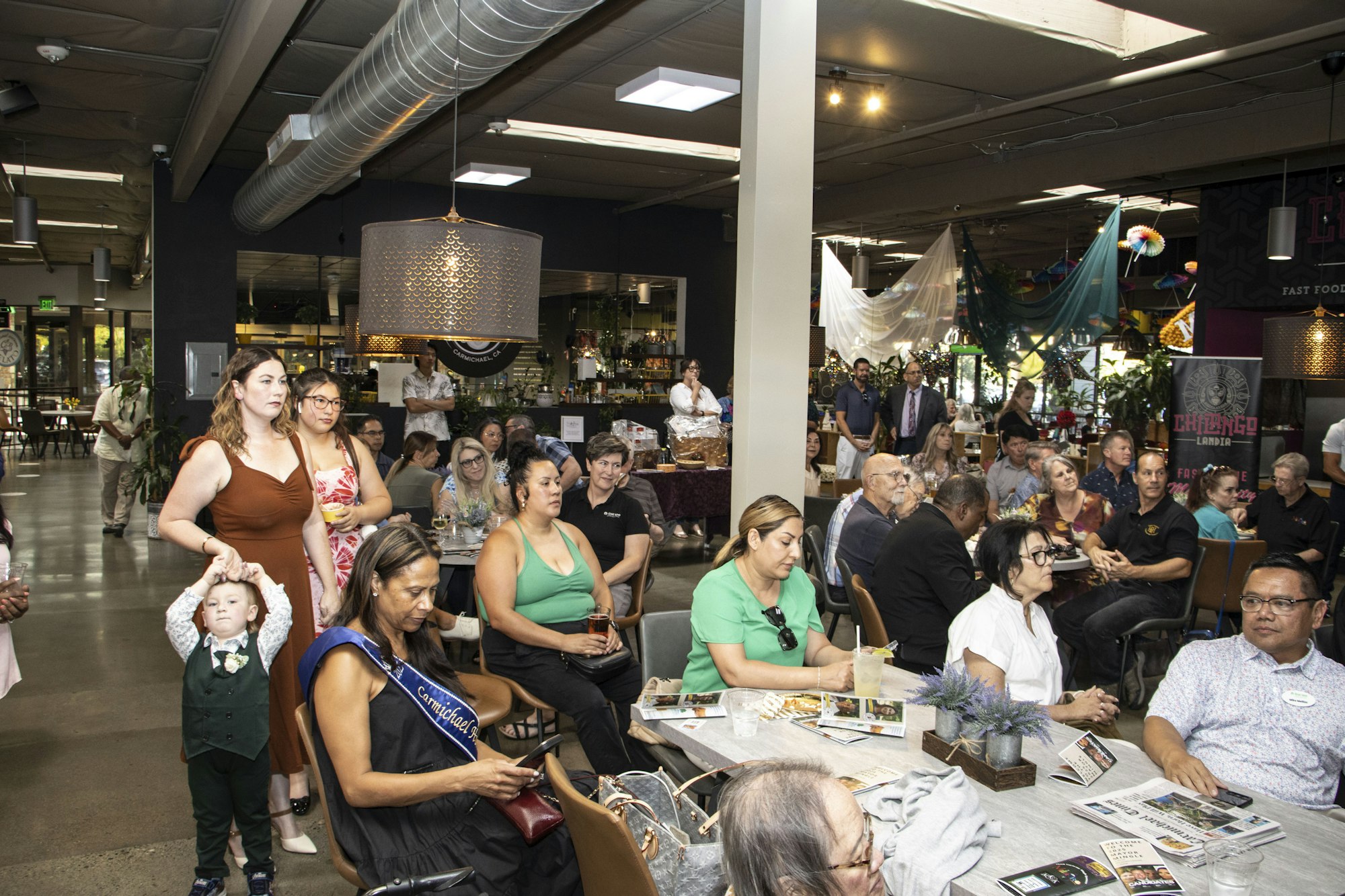 People gathered at an indoor event, sitting and standing, with food and decorations in the background.