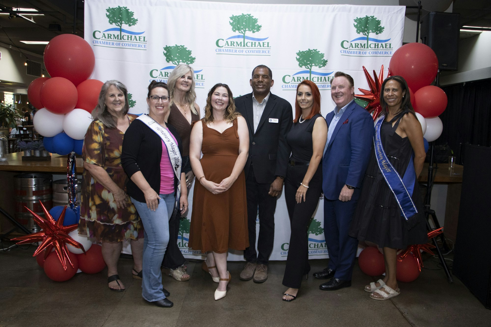 A group of people posing in front of a "Carmichael Chamber of Commerce" backdrop with red, white, and blue balloons.
