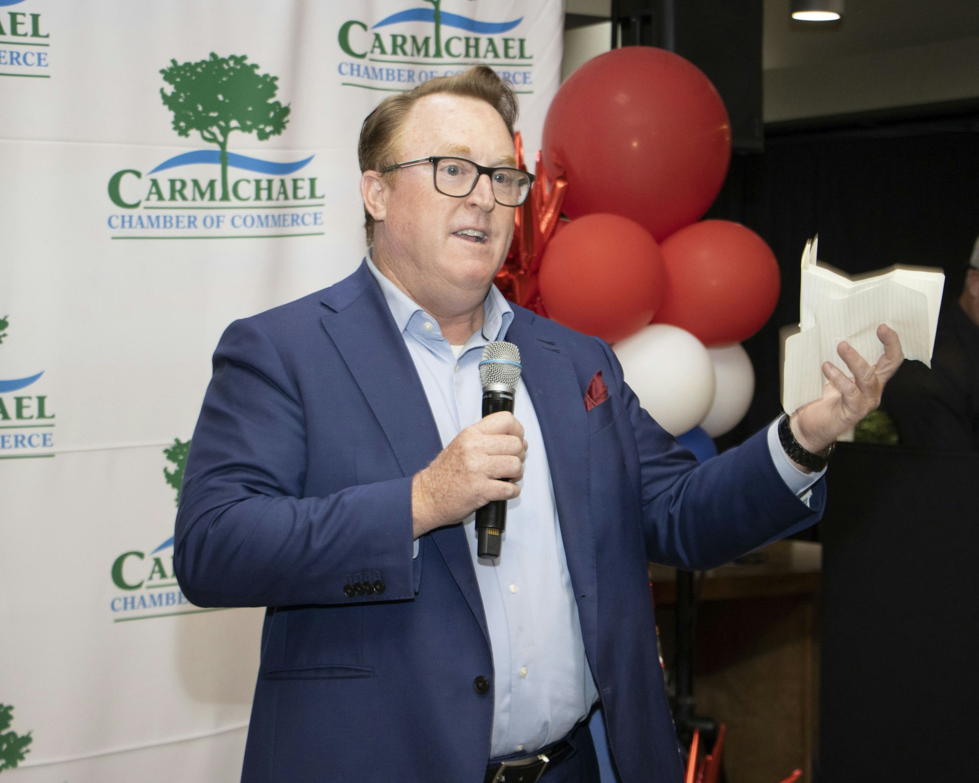 A man in a blue suit speaks into a microphone, holding a book. Red, white, and blue balloons are in the background.