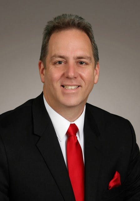 A person in a suit with a red tie and pocket square, smiling against a neutral background.