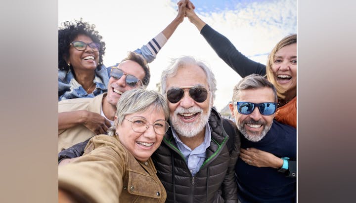 A joyful group of six people smiling, taking a selfie outdoors.