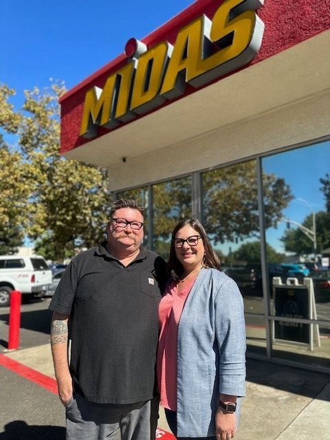 Two people standing in front of a Midas auto service shop on a sunny day.