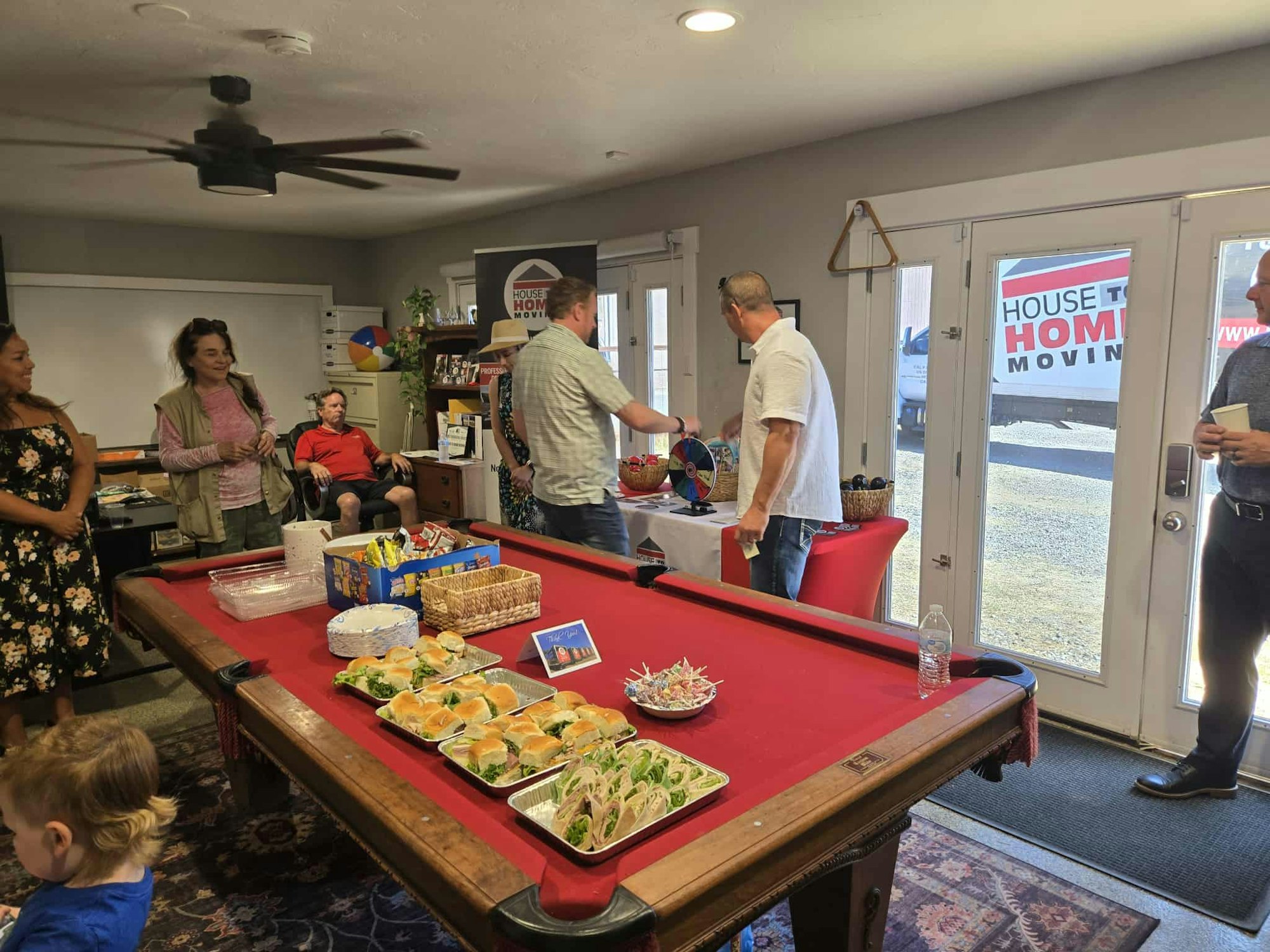 People gathered in a room with a pool table covered in food and snacks; a "House to Home Moving" sign is visible outside.