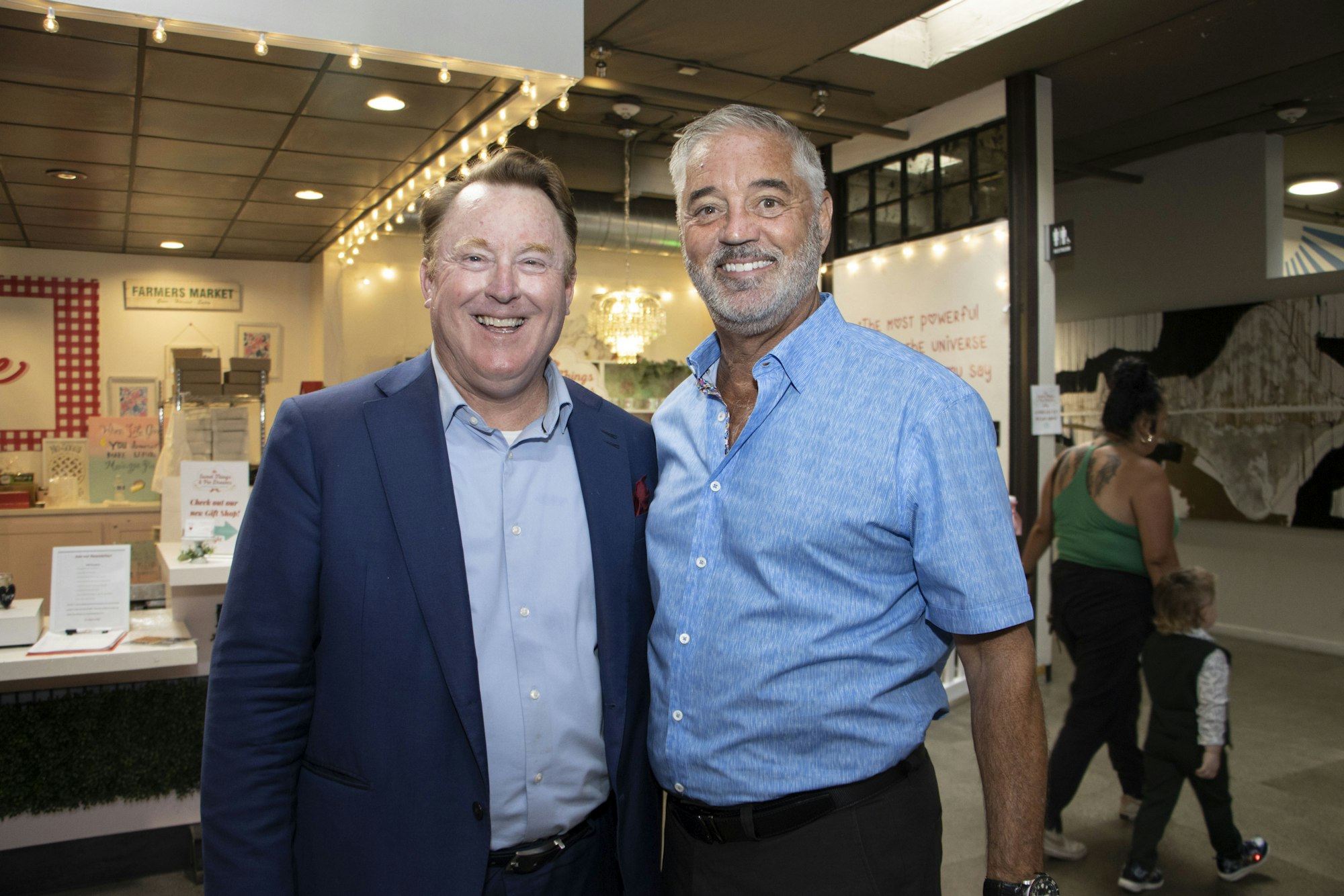 Two men smiling in a market setting with a "Farmer's Market" sign in the background.