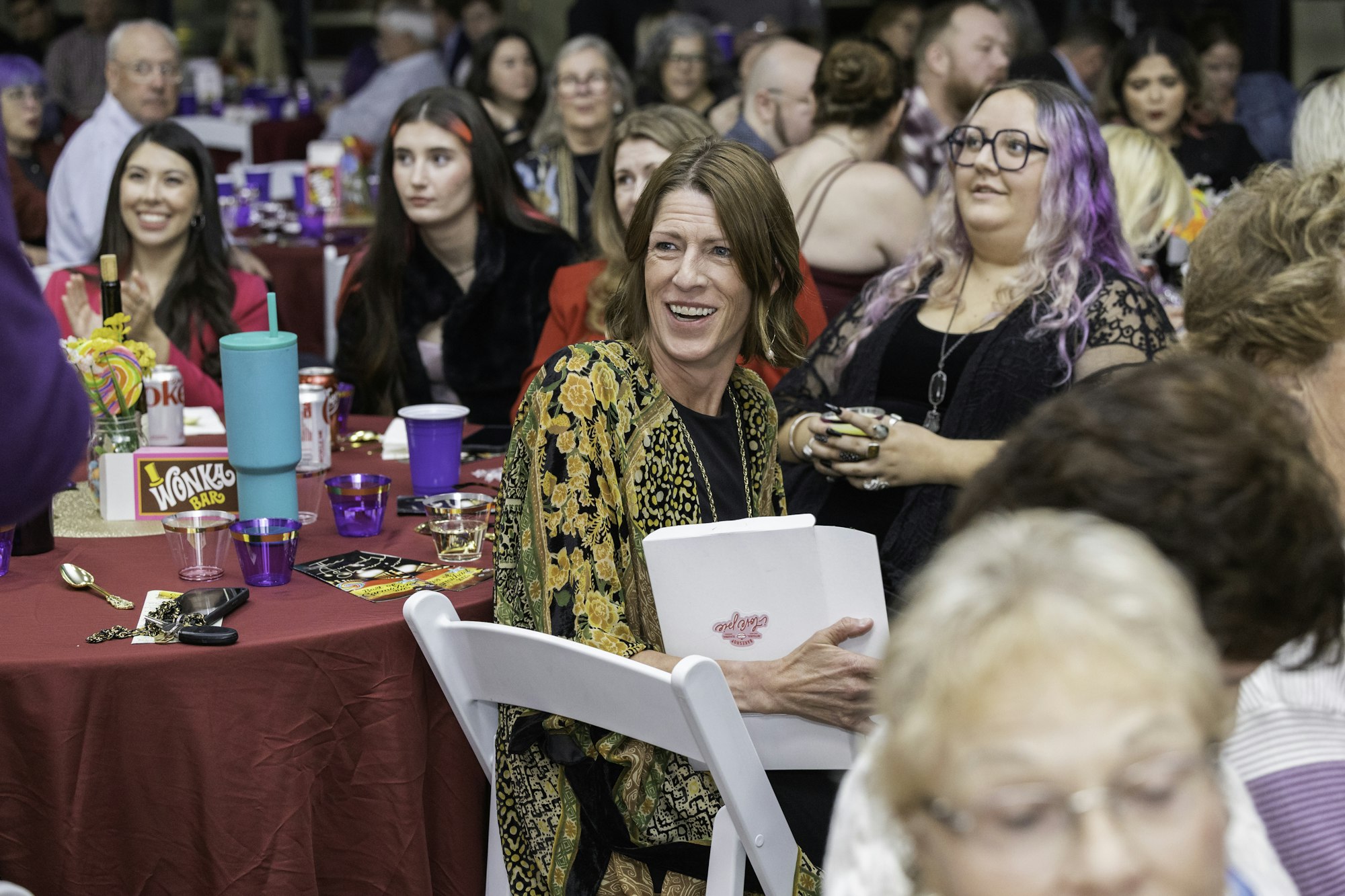 A group of people sitting at tables with drinks and decorations, a woman in a yellow floral outfit is smiling and holding a book.