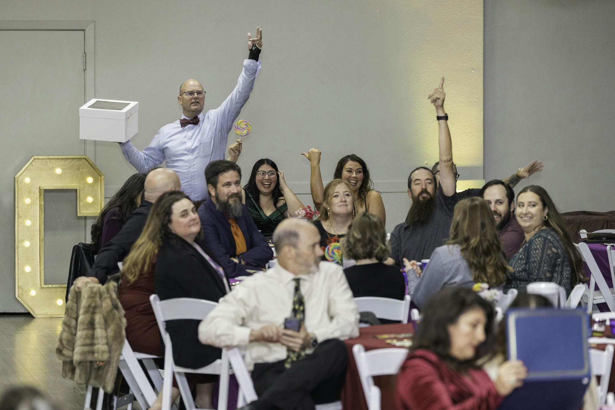 A group of people at a table, some raising their hands and smiling, with a man holding a white box.