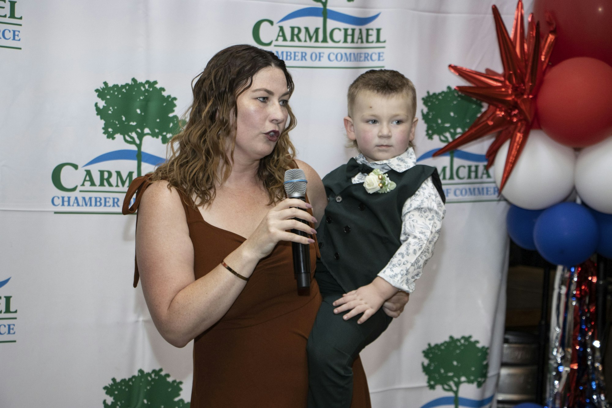 A woman holding a child speaks into a microphone at an event with "Carmichael Chamber of Commerce" signage and balloons in the background.