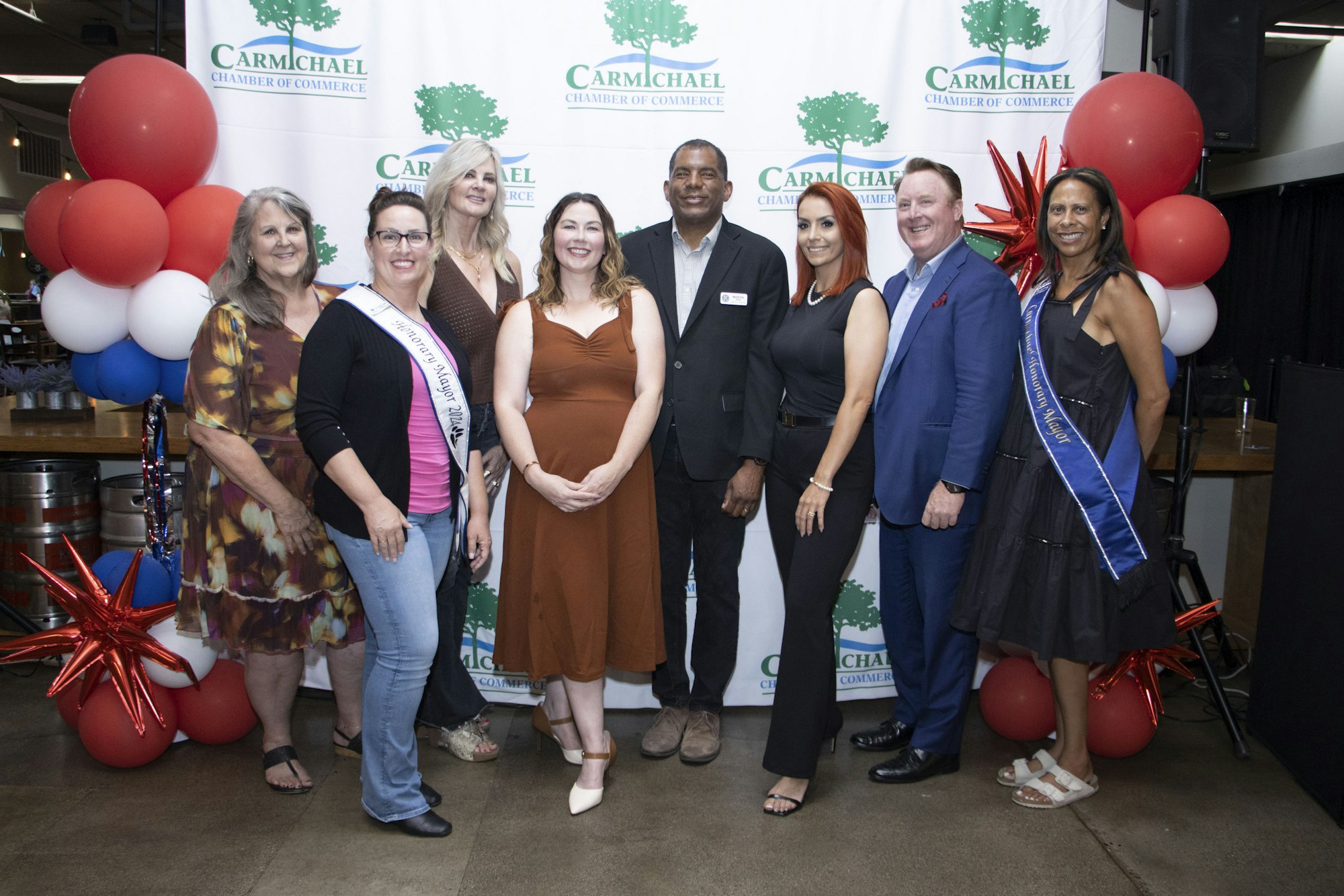 A group of people posing in front of a "Carmichael Chamber of Commerce" backdrop with festive balloons.