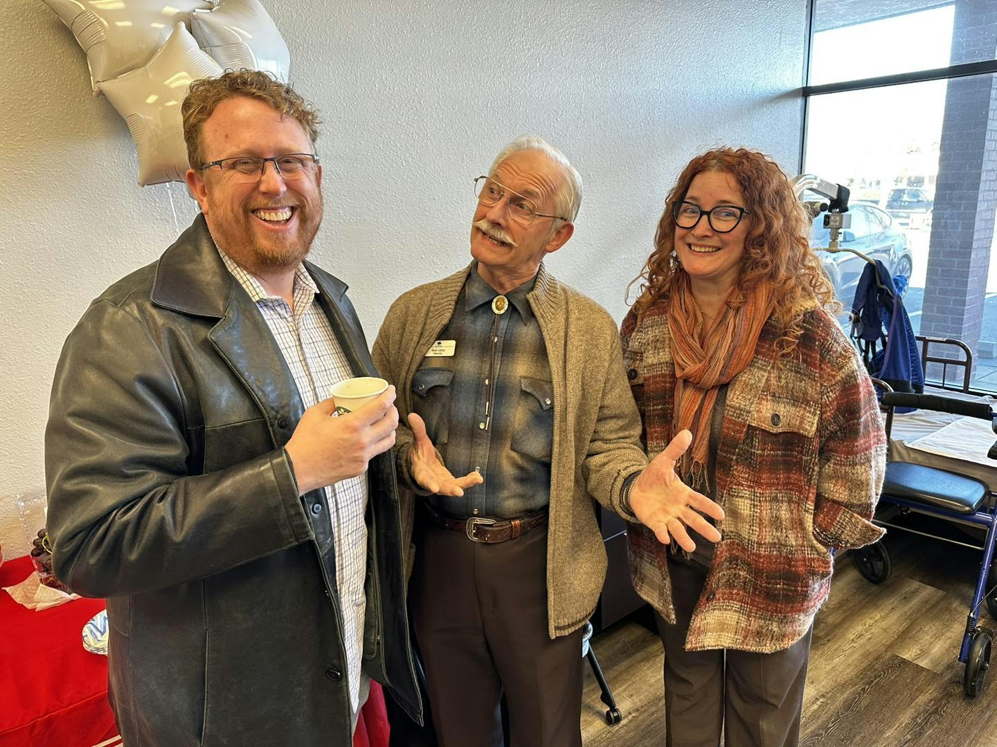 Three people laughing and chatting indoors, with balloons and a walker in the background.
