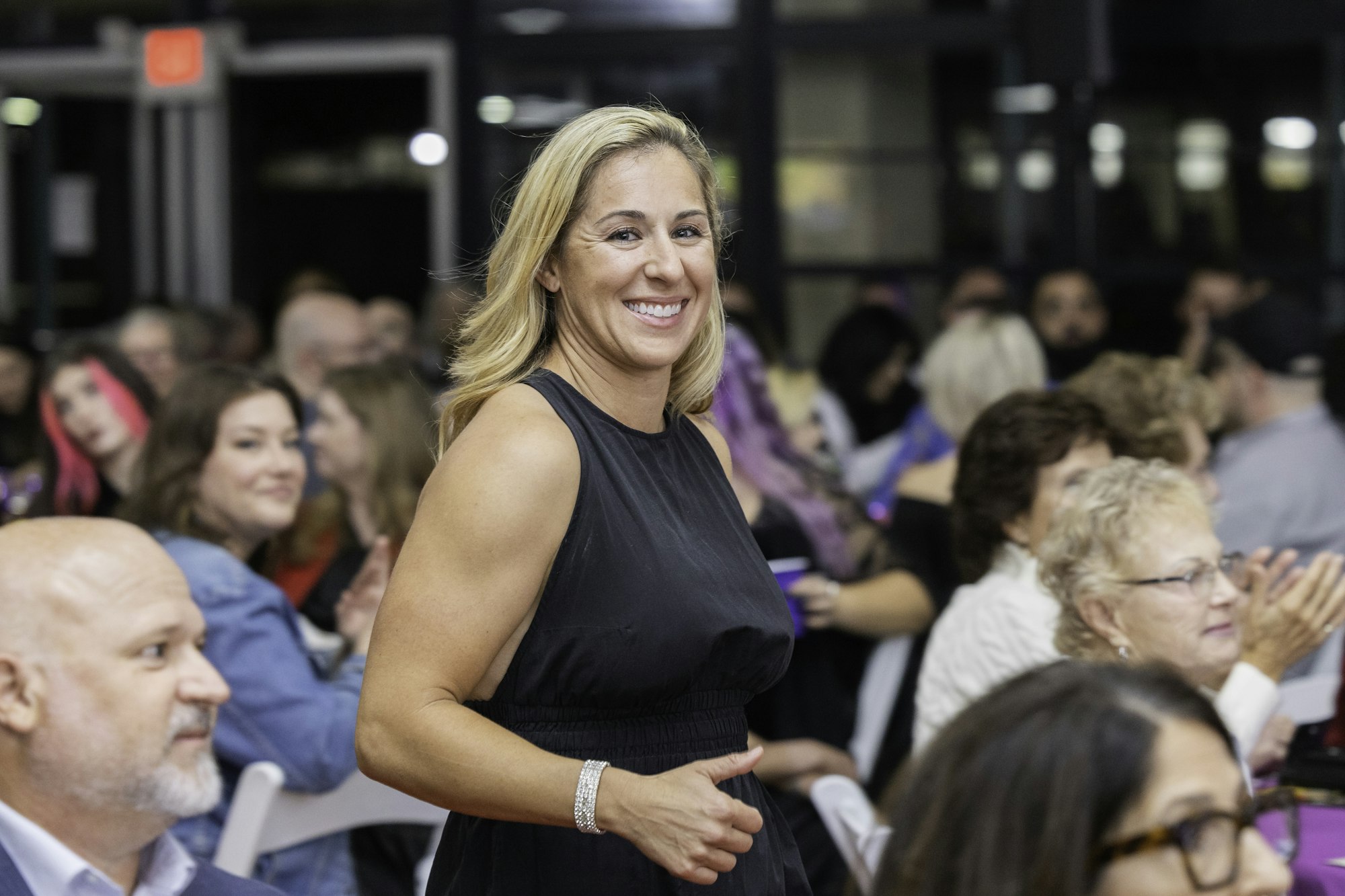 A woman in a black dress smiles at a crowded indoor event.