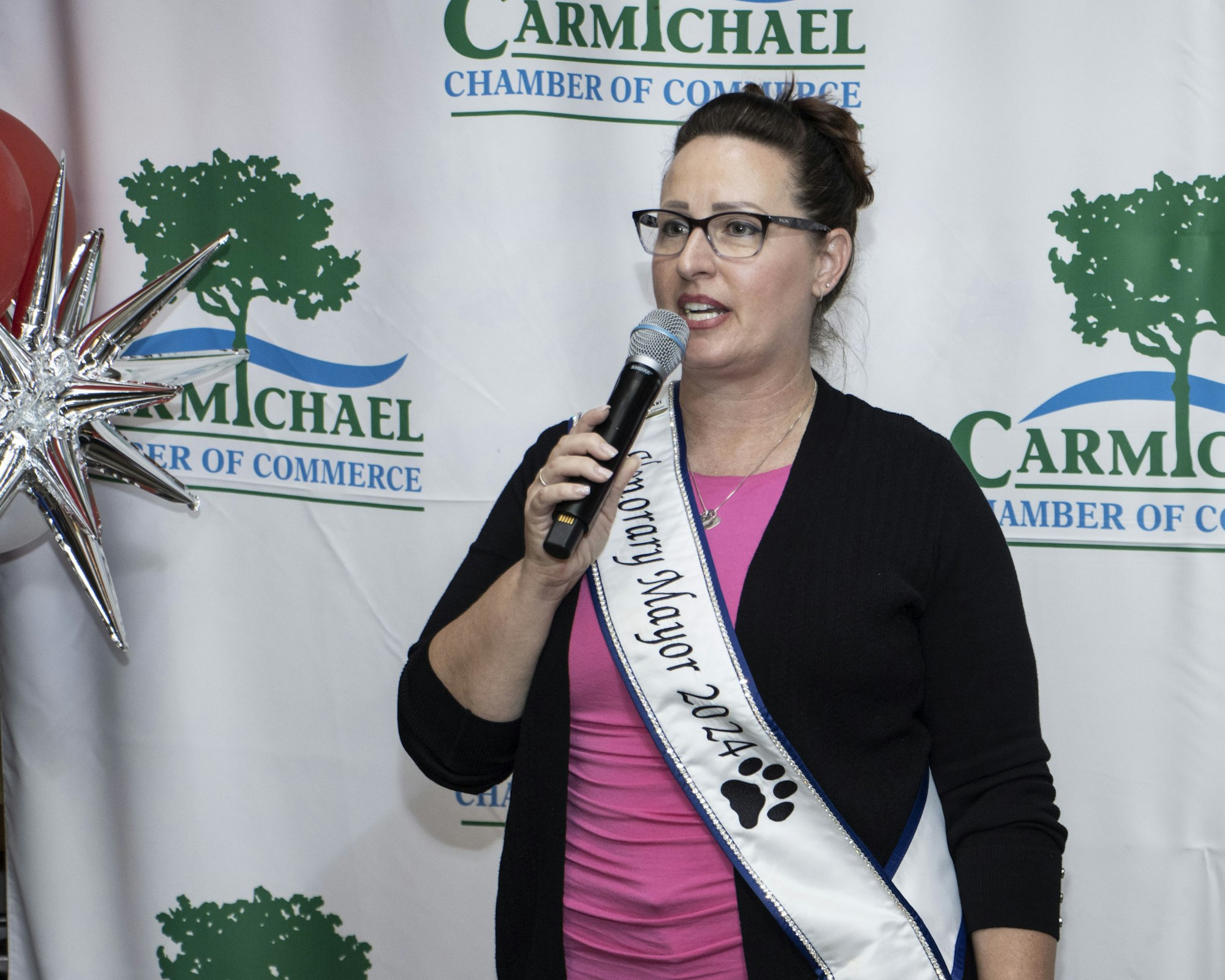 A person with a "Honorary Mayor 2024" sash speaking into a microphone at a Carmichael Chamber of Commerce event.