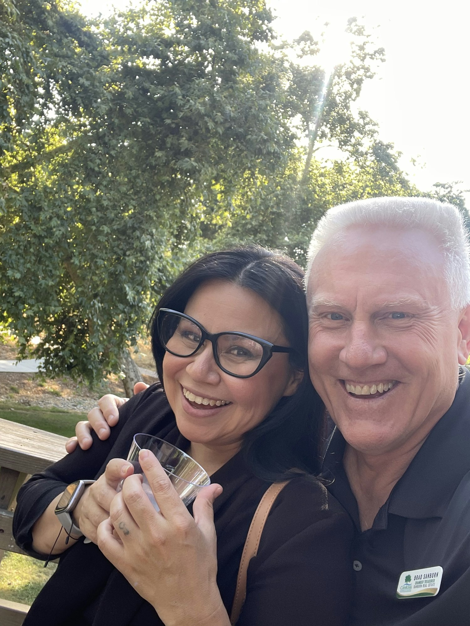 A smiling man and woman pose together outdoors, with trees in the background.