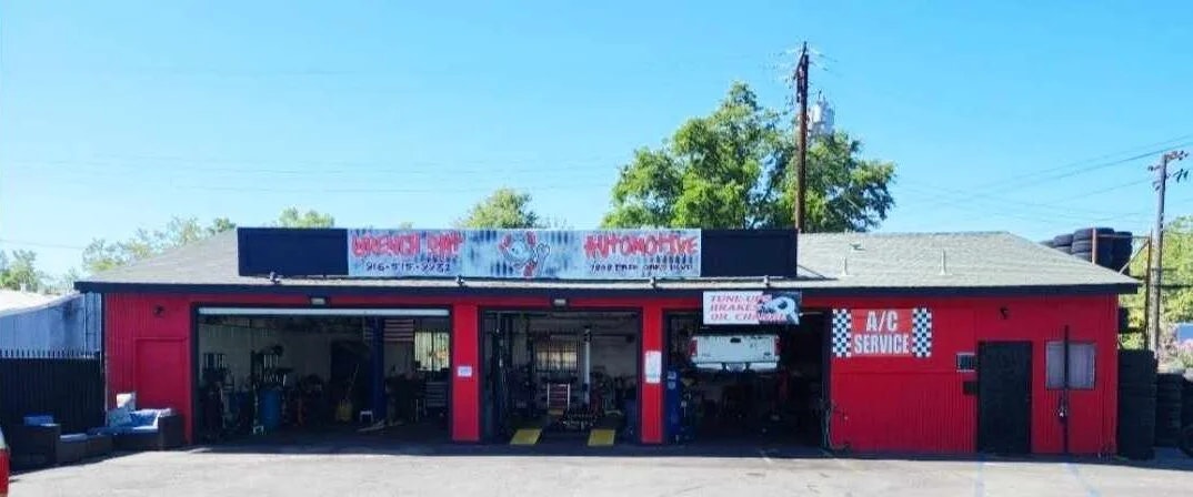 A red automotive repair shop with open garage bays, signs for A/C service, and trees in the background.