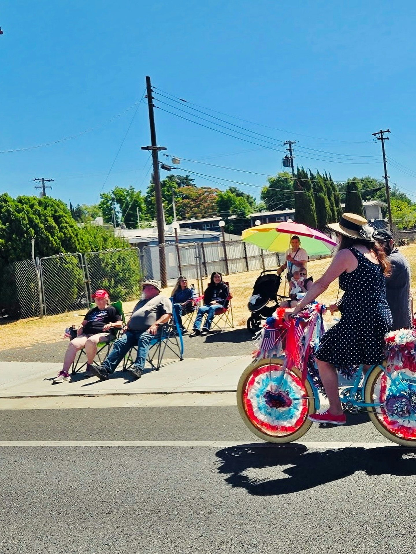 A person in a polka-dot dress rides a decorated bike past seated spectators on a sunny street.