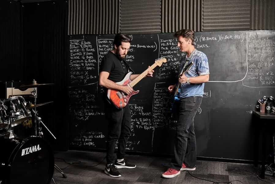 Two guitarists playing in a room with a drum set and music structure notes on a blackboard.
