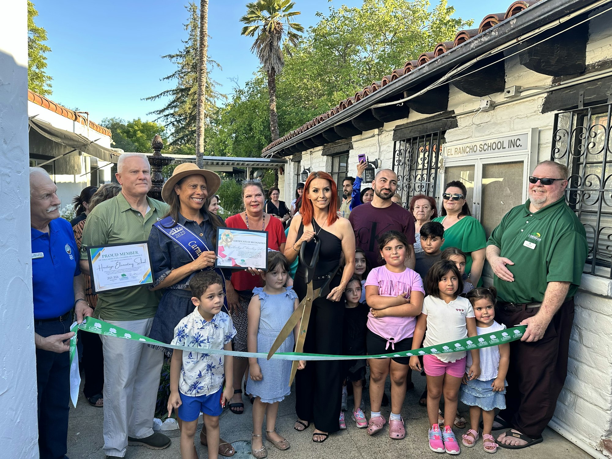A group of people, including children, at a ribbon-cutting ceremony for El Rancho School, with certificates and large scissors.