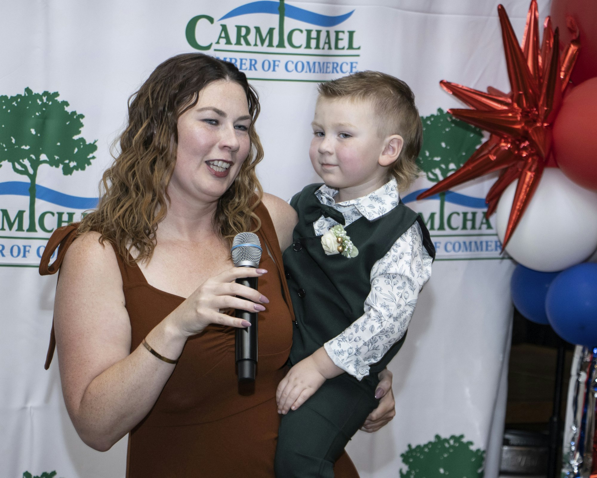 A woman holding a child, speaking into a microphone, in front of a "Carmichael Chamber of Commerce" backdrop with balloons.