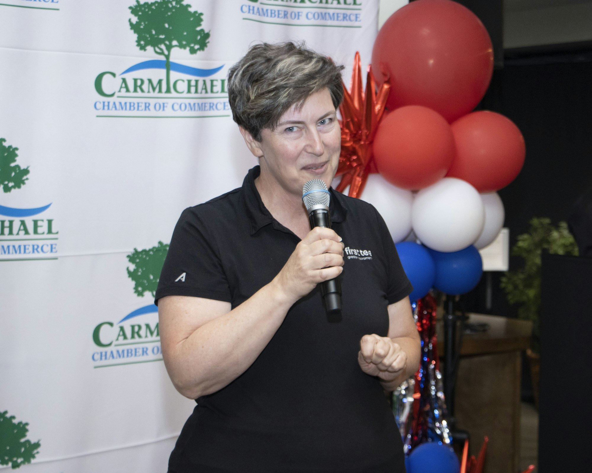 A person speaking into a microphone at a Carmichael Chamber of Commerce event, with a backdrop and red, white, and blue balloons.