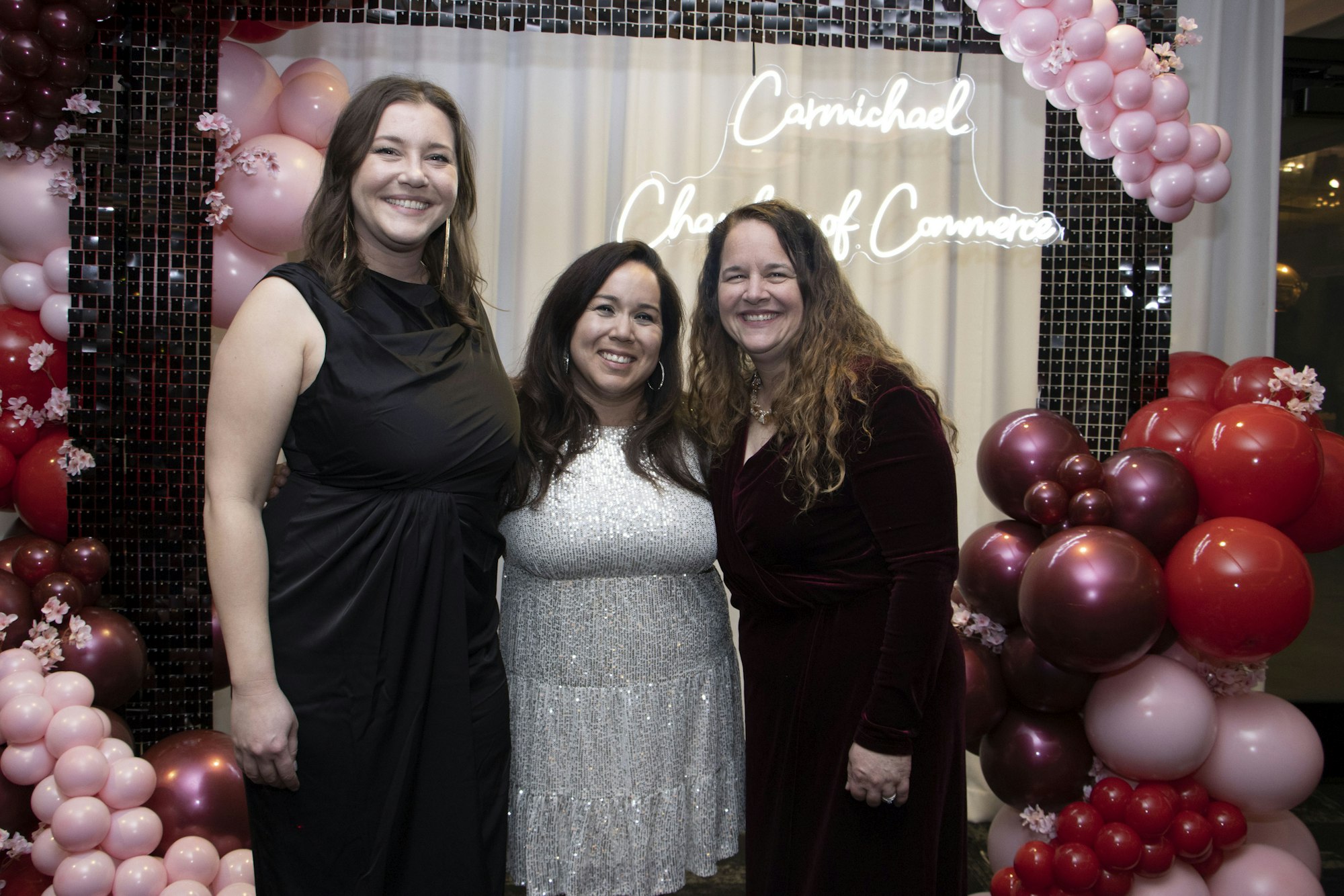 Three women posing together with a backdrop of pink and red balloons and a sign reading "Carmichael Chamber of Commerce."