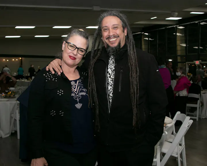 A woman and a man with distinctive hairstyles pose together at an indoor event with tables and guests in the background.