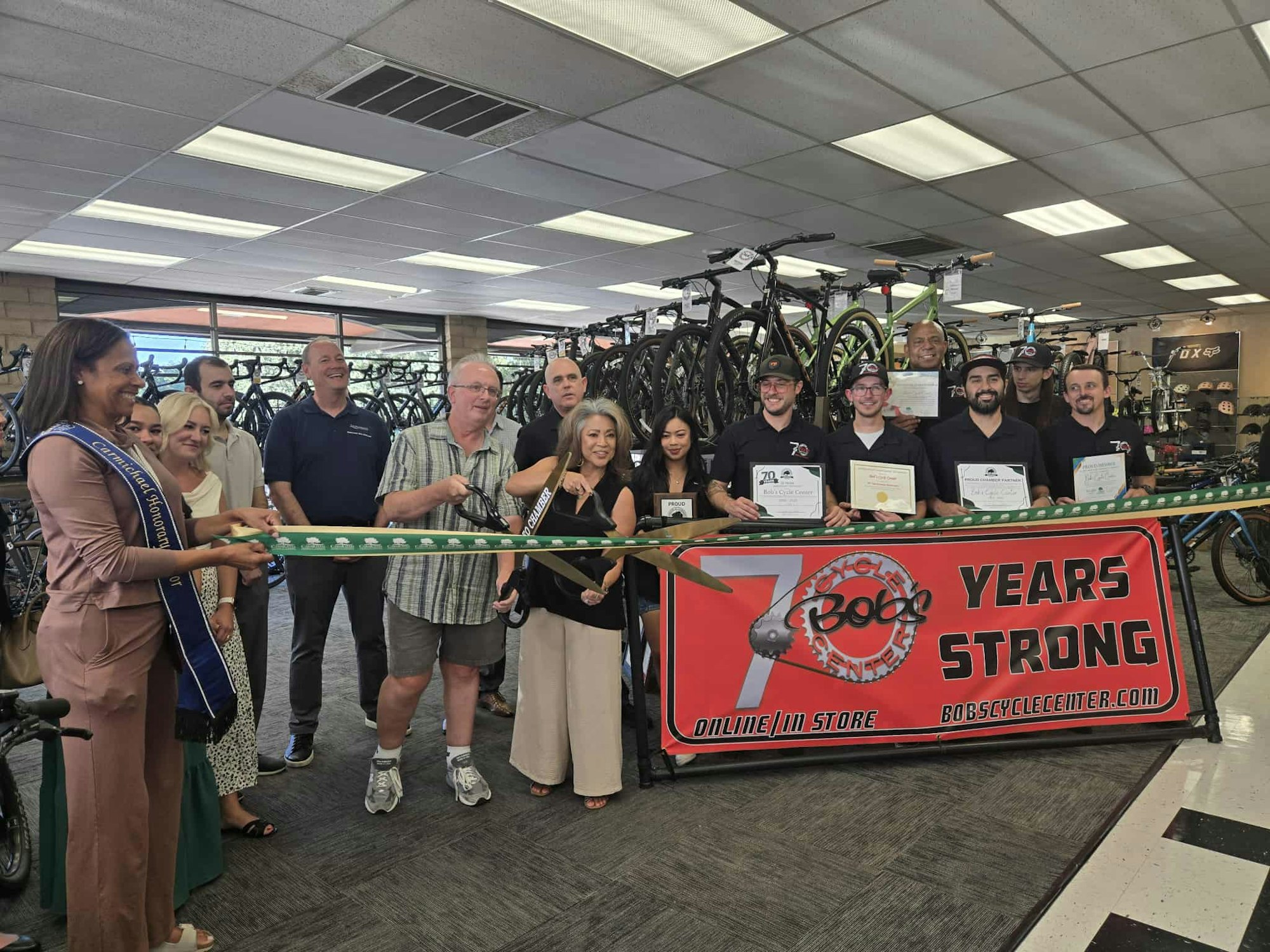 A group celebrates with a ribbon-cutting event in a bike shop, marking 70 years in business. People hold certificates, and bikes are displayed.