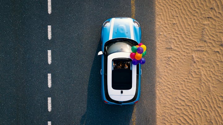 Aerial view of a blue car with a white roof and colorful balloons, driving along a road adjacent to a sandy terrain.