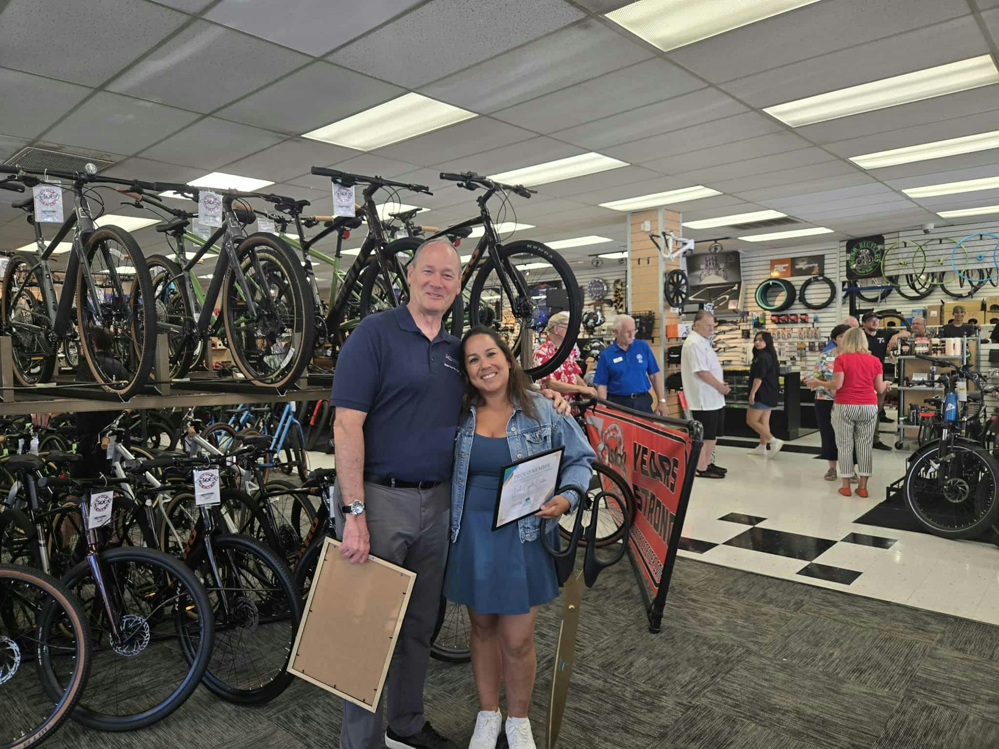 Two people posing in a bike shop, holding certificates and a large pair of scissors. More people and bicycles in the background.