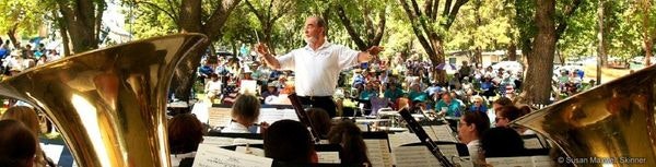 A conductor leads an outdoor orchestra performance; audience in the background, large brass instruments in the foreground.