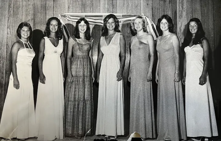 A group of seven women in elegant dresses posing together in a vintage setting, likely for a special event or photo shoot.