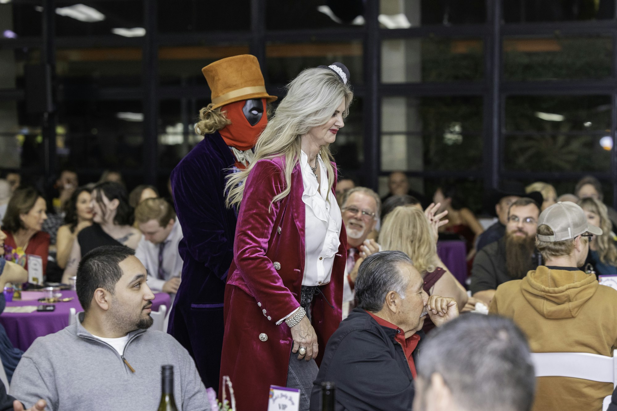 A costumed individual in a hat and mask stands behind a person in pink walking through a crowded room.