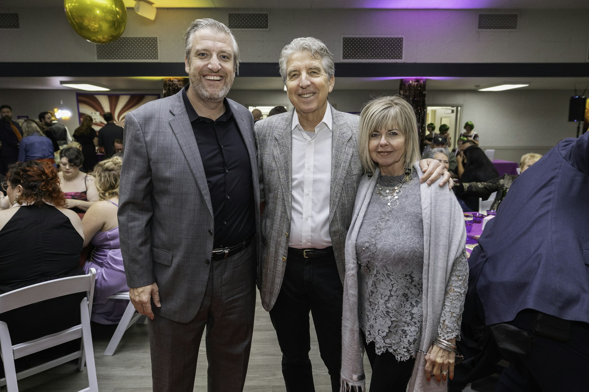 Three people posing at an indoor event, with a crowd in the background and a large gold balloon above.