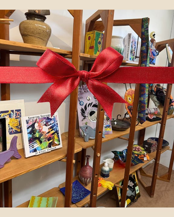 A display of colorful art items and pottery on wooden shelving, adorned with a large red bow.