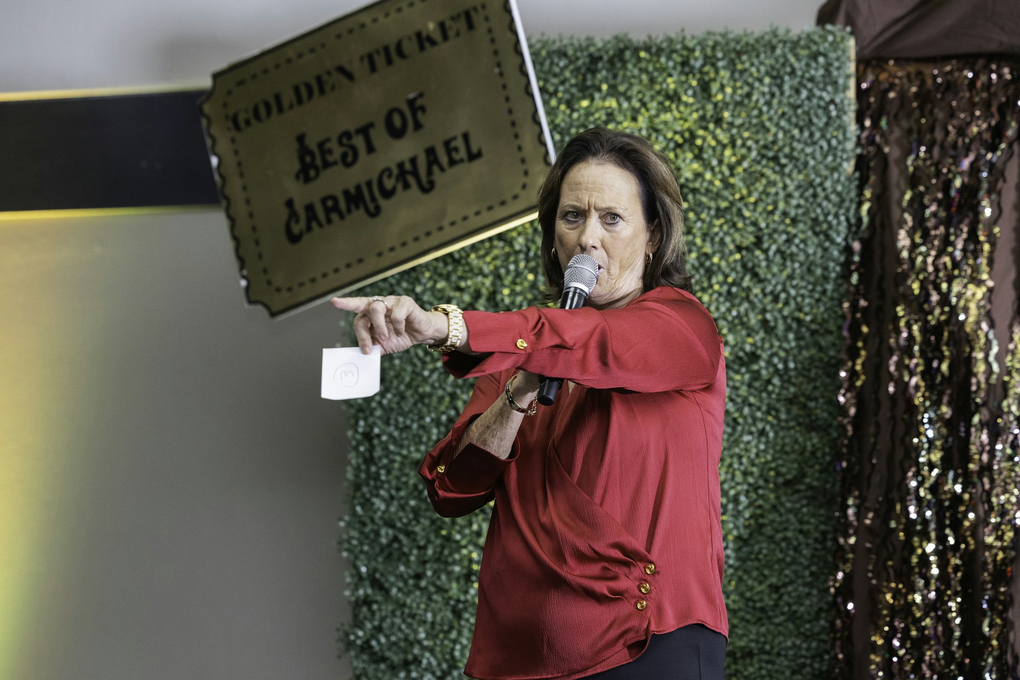 Person in red speaking with a microphone, pointing with a paper, "Golden Ticket: Best of Carmichael" sign in the background.