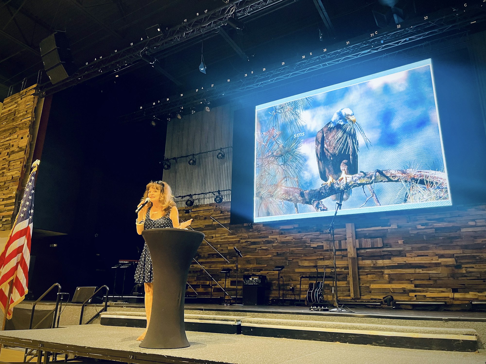 A person speaks on stage with a microphone, standing next to an American flag, in front of a large screen displaying an eagle.
