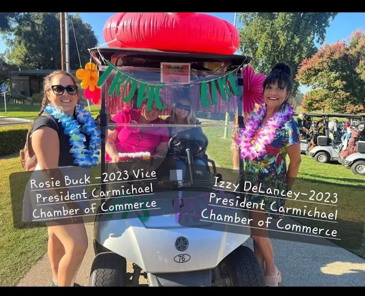 Two women in tropical attire pose with a decorated golf cart at an event. Text highlights their roles in the Carmichael Chamber of Commerce.