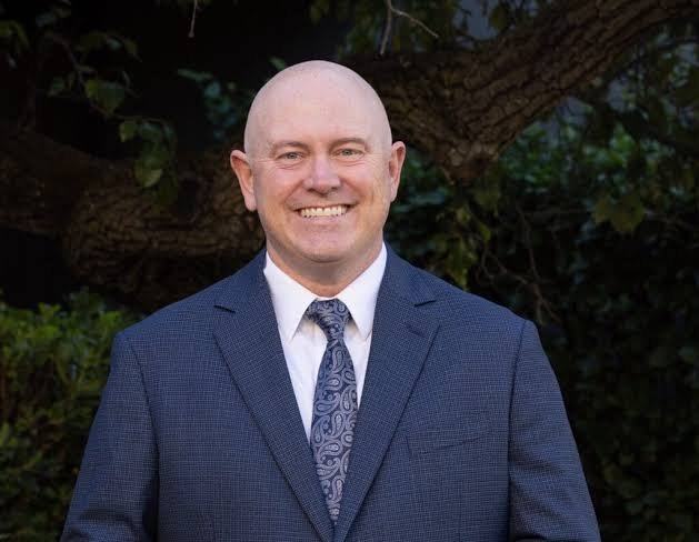 A smiling bald man in a suit and tie stands outdoors, with a tree and greenery in the background.