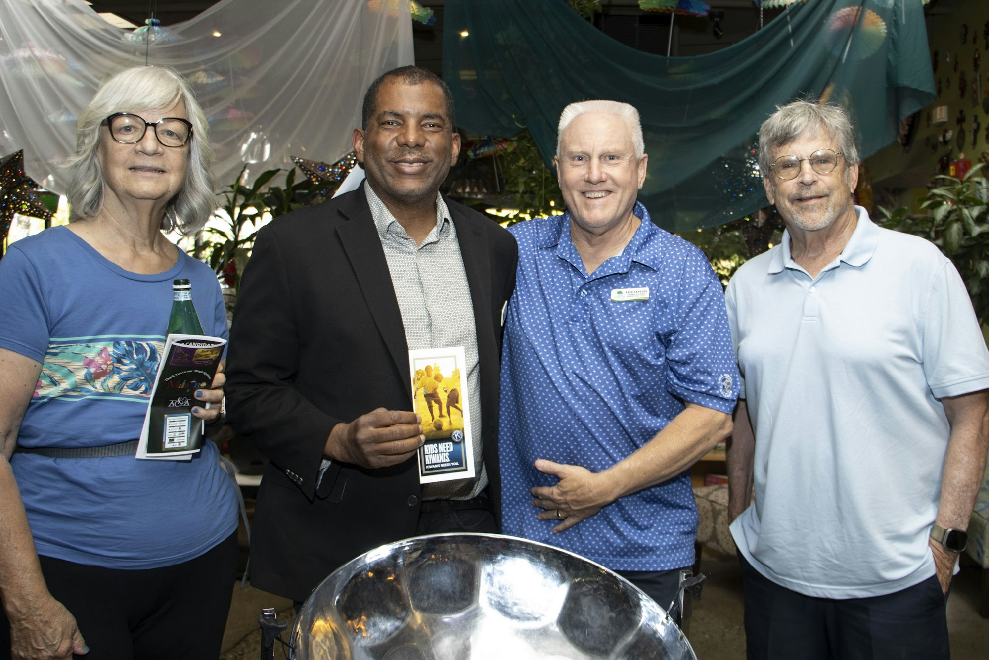 Four people smiling, standing together indoors, two holding flyers, near a steel drum.