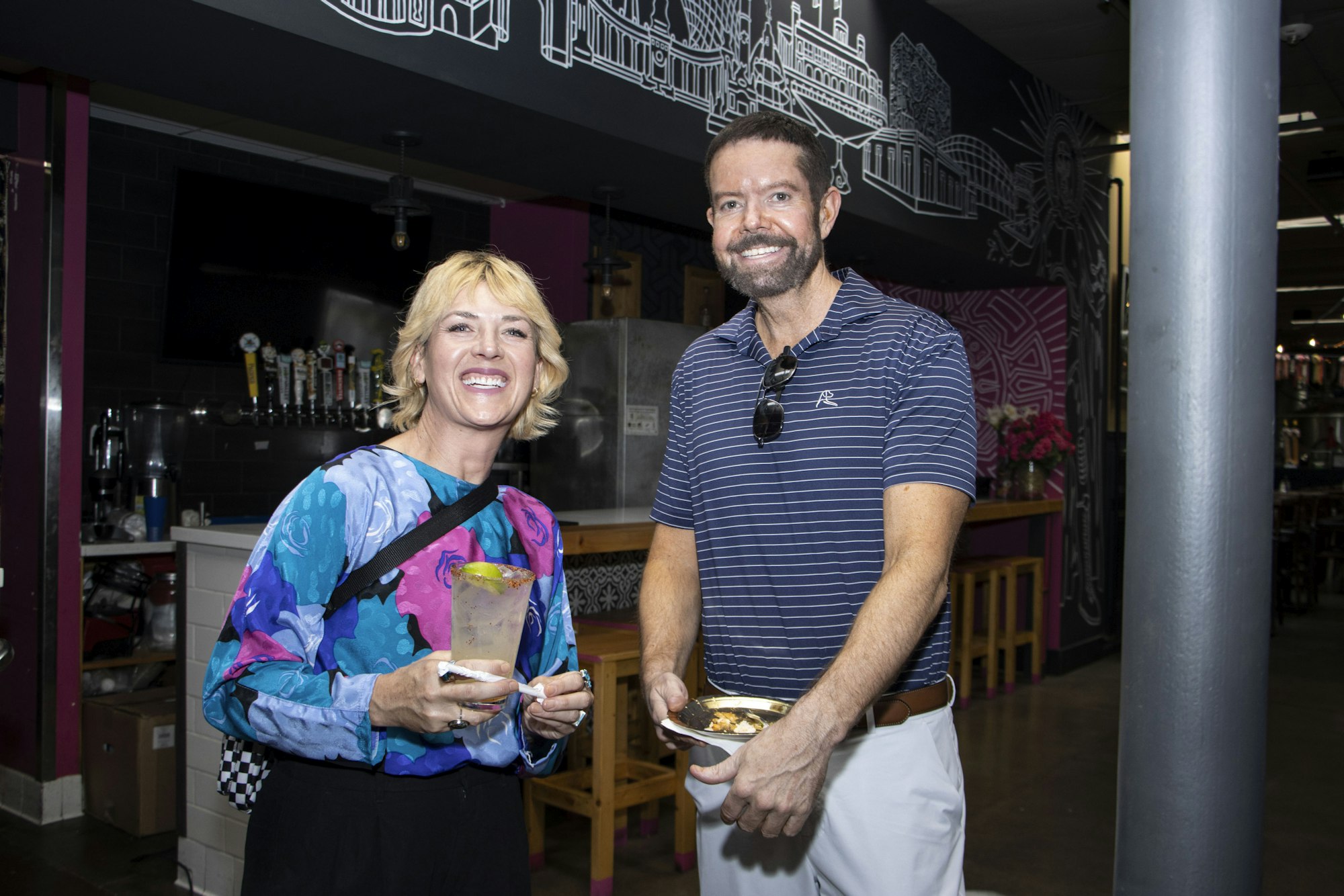 Two people smiling indoors, one holding a drink, the other holding a plate of food, with a bar and artwork in the background.