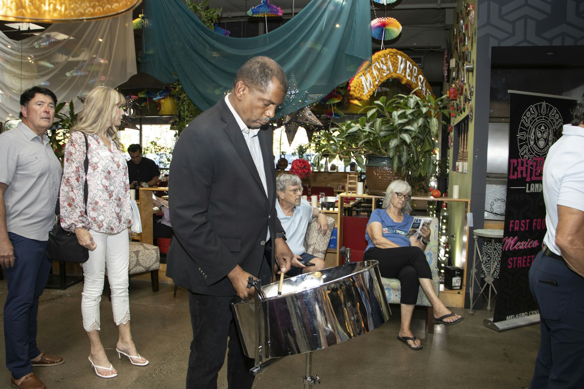 People in a decorated area, one playing a steel drum, others sitting or standing, with a colorful backdrop and plants.