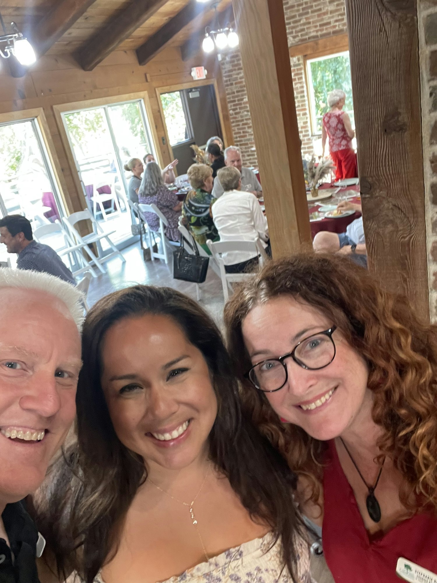 Three people are smiling for a selfie in a room with a gathering in the background.