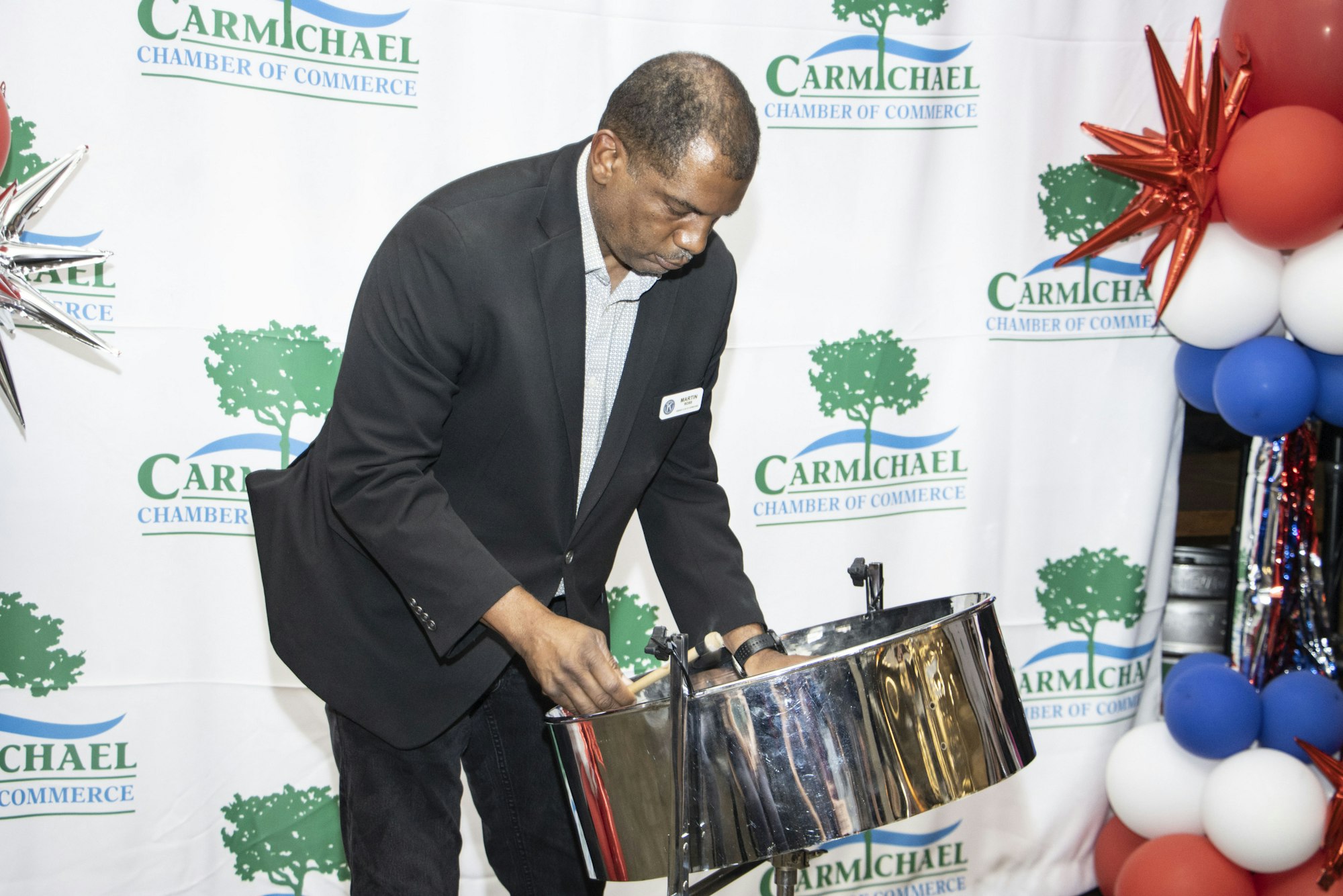 A person in a black suit plays a steel drum at a Chamber of Commerce event with balloons and a branded backdrop.