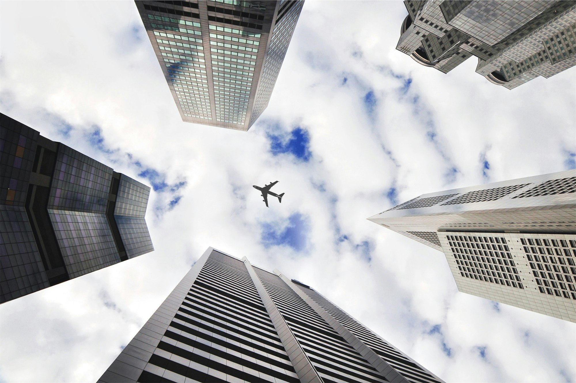 Looking up at skyscrapers with an airplane flying in a cloudy sky.
