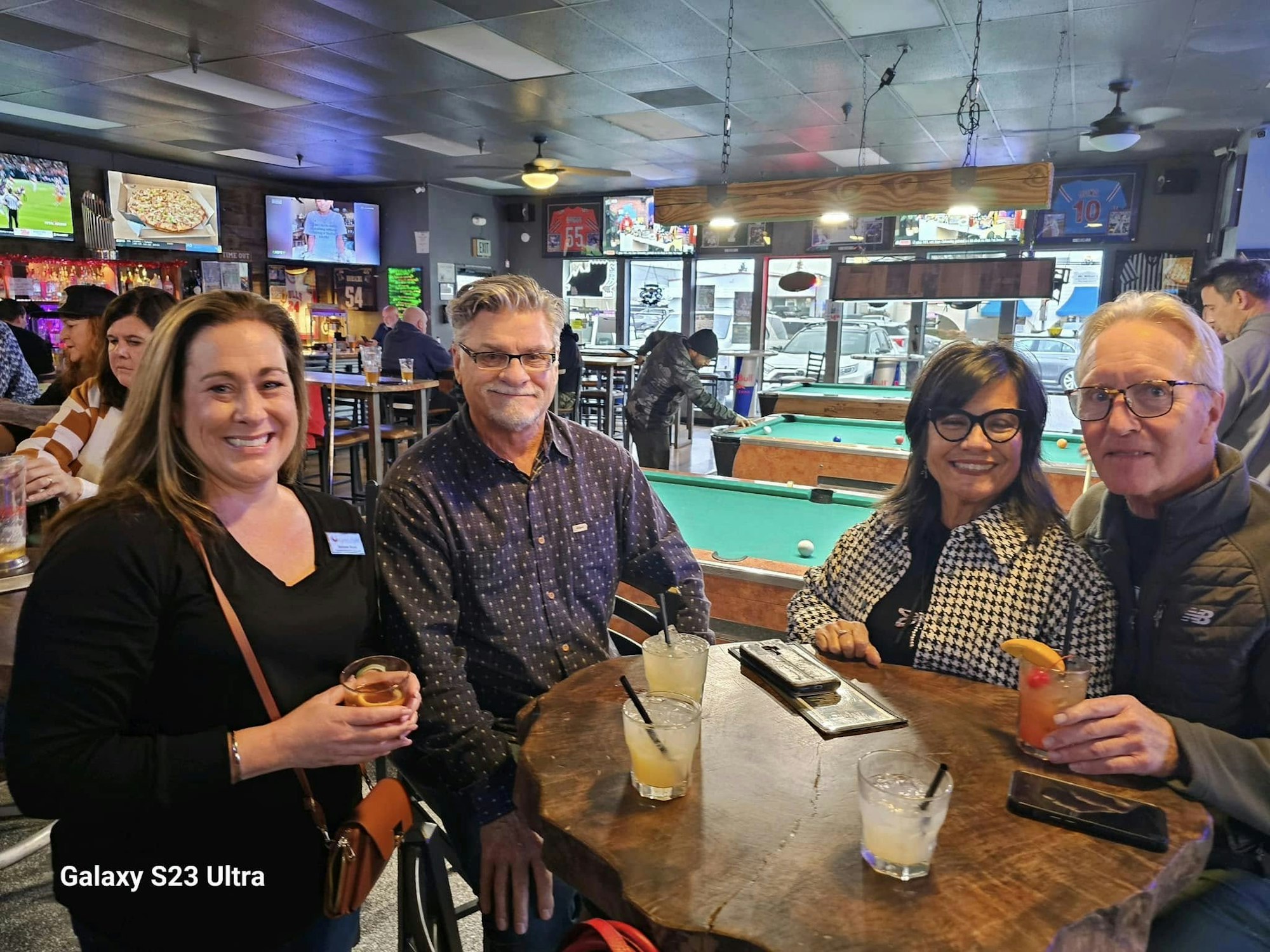 Four people smiling at a bar table, holding drinks. Pool tables and TV screens in the background.