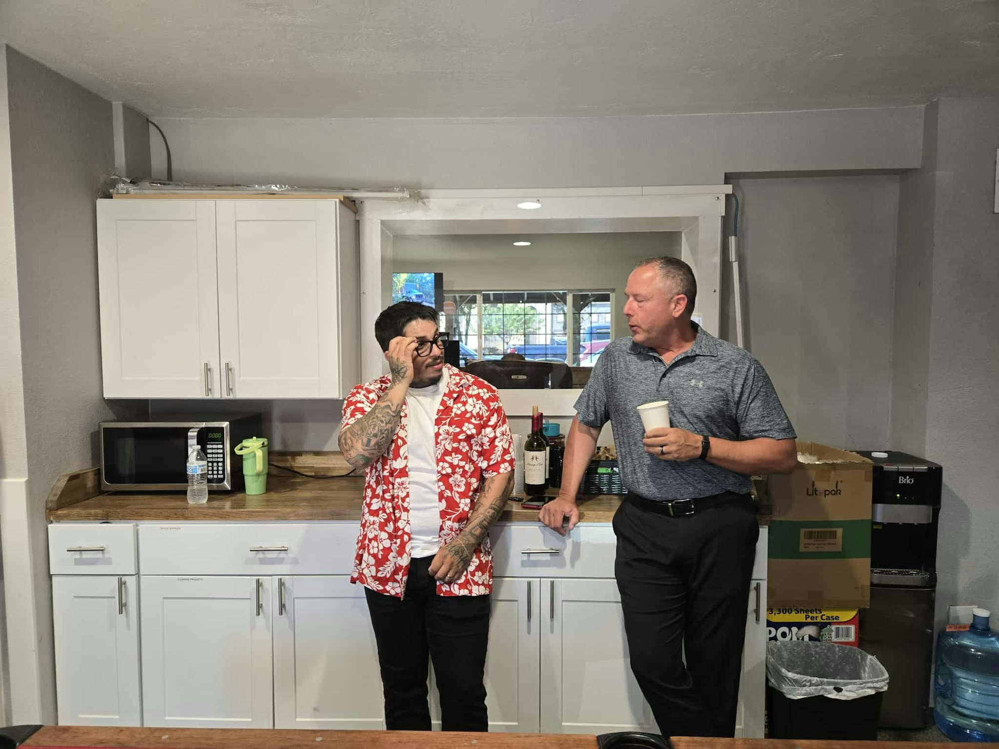 Two men talking in a kitchen area, one in a red floral shirt. There are cabinets, a microwave, and a water cooler in the background.